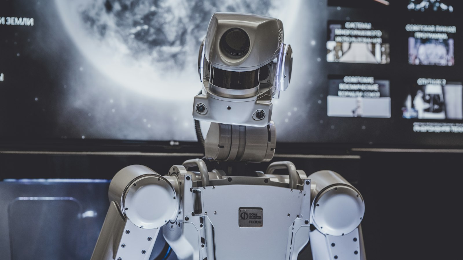 A white and grey humanoid robot standing in front of a wall of screens